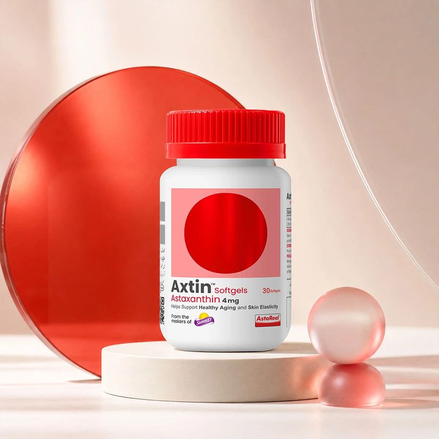 A white bottle of Axtin Softgels Astaxanthin 4mg with a red cap, displayed on a beige pedestal with a large red circular prop and pink spheres in a studio setting.
