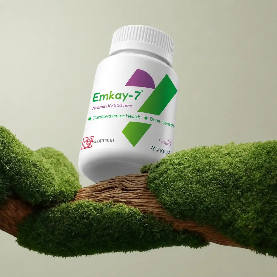 A white plastic bottle of Emkay-7 Vitamin K3 200 mcg stands on a mossy branch against a light beige background. The label features green and purple geometric accents and text indicating cardiovascular and bone health benefits.