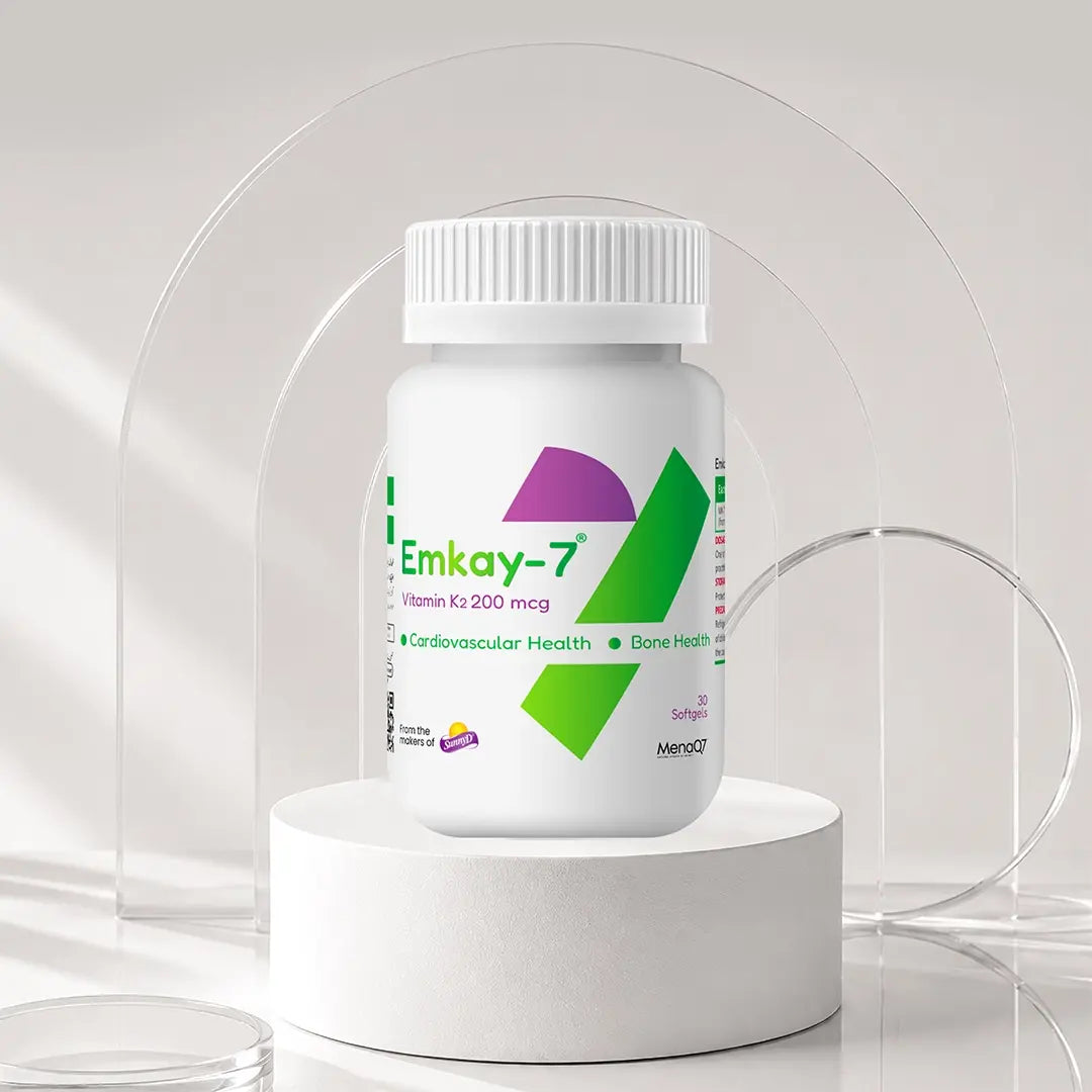 A white plastic bottle of Emkay-7 Vitamin K2 200 mcg capsules, displayed on a white cylindrical pedestal with a clear acrylic arch background.