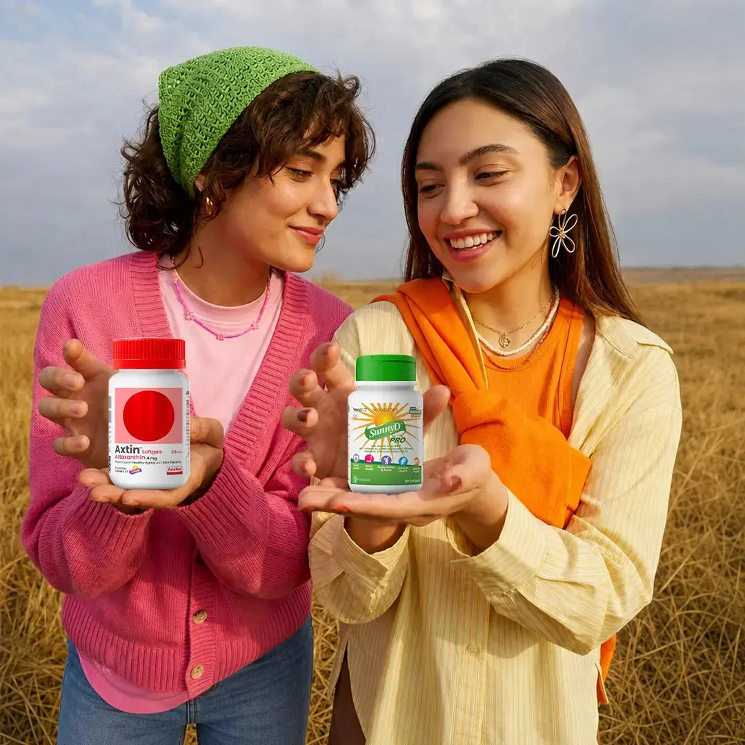 Two women standing in a field of dry grass, each holding a supplement bottle. The woman on the left wears a green beanie, a pink cardigan, and blue jeans, holding a white bottle with a red lid labeled 'Latina'.
