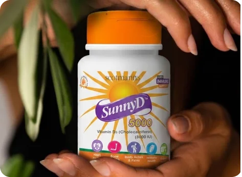 A person holds a bottle of Scottmanns SunnyD 5000 Vitamin D3 supplements. The white bottle features a bright orange cap and a label with a yellow sun graphic and purple brand banner.
