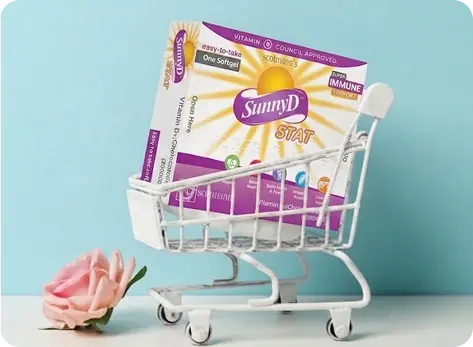 A miniature white shopping cart holding a box of SunnyD STAT vitamin D supplement, set against a light blue background with a pink rose on the floor.