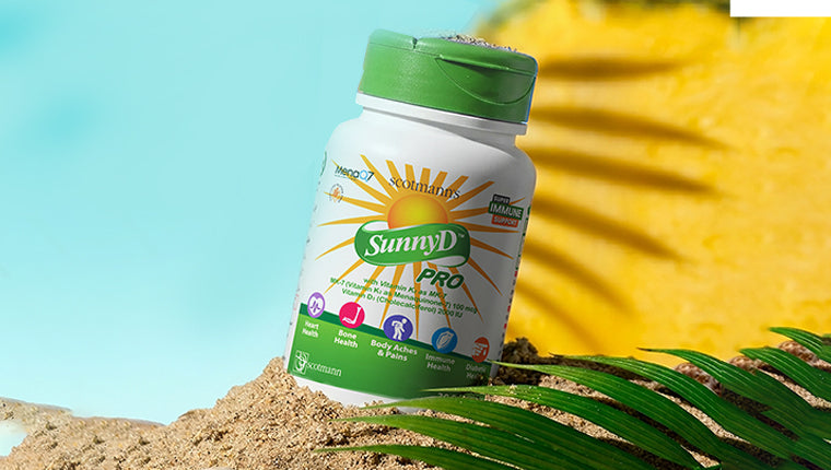 Scotmann's SunnyD Pro vitamin D3 and K2 MK-7 softgel supplement bottle on sand with a tropical palm leaf and blue and yellow background.