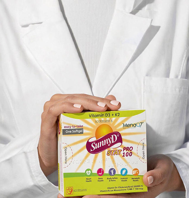 A person wearing a white lab coat holds a box of SunnyD Star Pro 100 Vitamin D3 + K2 softgels. The box features a yellow and white design with a prominent purple SunnyD logo.