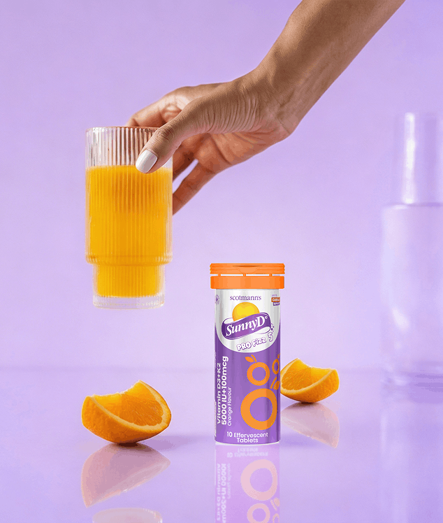 Hand holding a glass of orange juice next to SunnyD drink mix packaging on a purple background